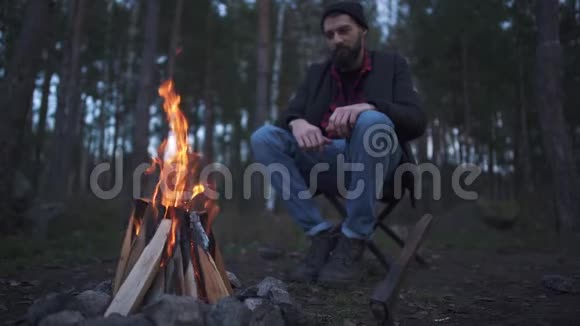 坐在火炉旁的椅子上的一个留胡子的年轻人森林里篝火旁的预告片视频的预览图