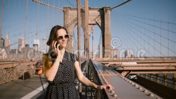 快乐的欧洲女商人戴着复古太阳镜在电话里交谈在布鲁克林大桥4K吹着长发视频的预览图