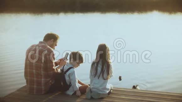 父亲和两个孩子坐在湖边码头上欧洲男人带着孩子用智能手机拍摄水鸟4K视频的预览图