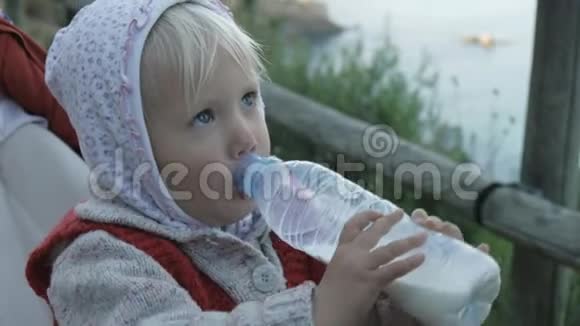穿着帽衫和红色针织背心的小女孩在婴儿车上坐着塑料瓶喝牛奶或奶制品视频的预览图