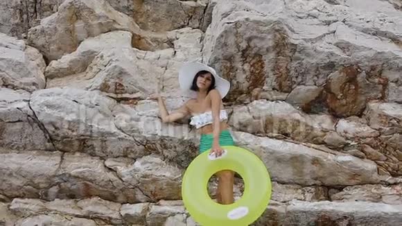 穿着泳衣和帽子的漂亮晒黑的女孩在靠近悬崖的相机上摆出一个可充气的黄色圆圈或视频的预览图
