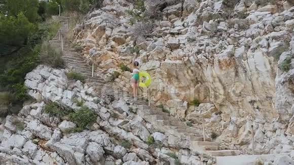 苗条的女孩穿着白色的绿色泳衣和大帽子走在靠近高岩石的台阶上带着充气来到海滩上视频的预览图