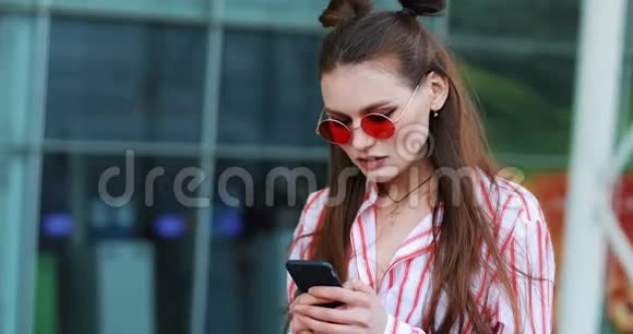 年轻女子站在一个购物中心附近打电话穿粉色太阳镜的女人在阳光明媚的日子写博客视频的预览图