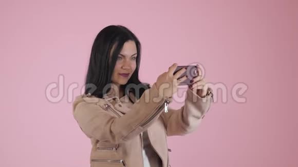 微笑快乐的黑发女人穿着粉红色夹克在智能手机上自拍背景五颜六色视频的预览图