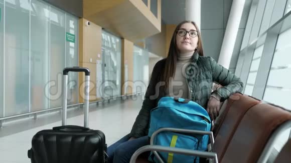 戴眼镜的年轻疲惫的女人正坐在大厅里的机场等待她的航班视频的预览图