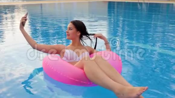 女孩在手机上被拍到躺在度假胜地的游泳池里的粉红色充气戒指上视频的预览图