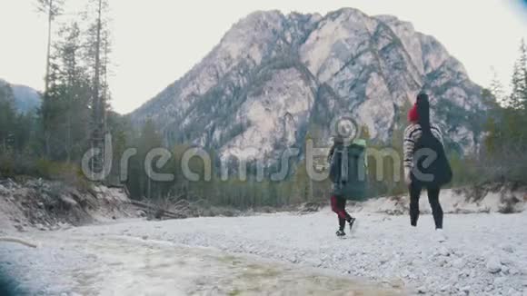两个年轻的女人在岩石河沿岸散步背着背包和吉他在森林的背景下视频的预览图