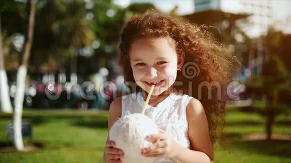 小白种人卷发的小女孩穿着白色连衣裙喝着椰子视频的预览图