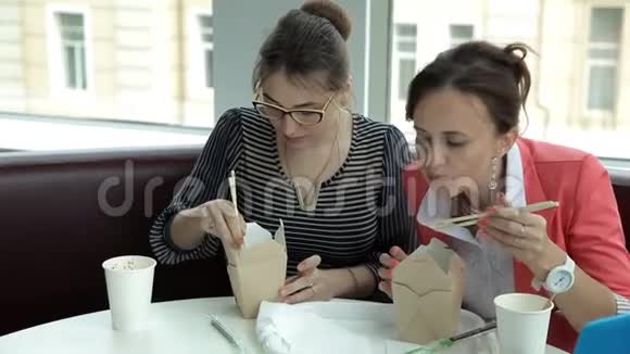 午餐时两个年轻的商务女孩坐在盒子里吃美味的面条喝咖啡工作午餐视频的预览图