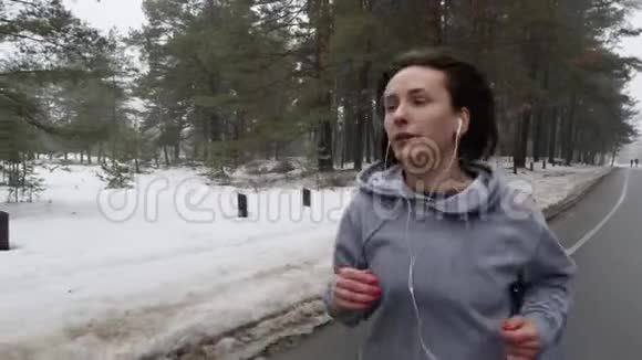 年轻迷人的白种人女孩在冬天带着耳机在雪地公园跑步关闭前方跟随射击慢动作视频的预览图