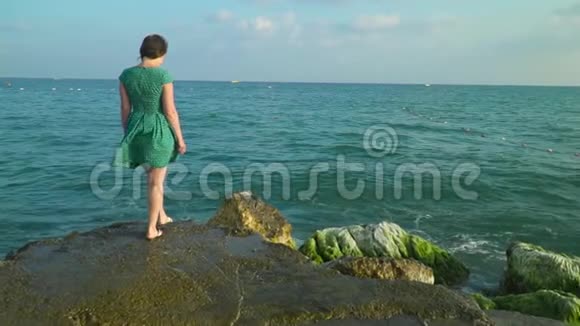 穿着绿色短裙的女人在潮湿的岩石上散步阳光下溅起海水慢动作视频的预览图