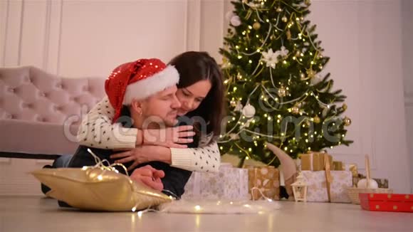 美丽的年轻情侣穿着圣诞老人帽躺在壁炉旁边的地板上装饰得很漂亮视频的预览图