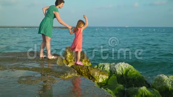 妈妈和女儿站在海水中潮湿的岩石上周围有飞溅的波浪玩得很开心慢动作视频的预览图