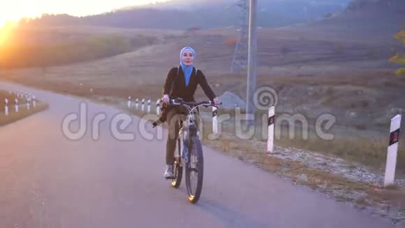 穆斯林妇女戴头巾背着背包骑着自行车在路上夕阳西下视频的预览图