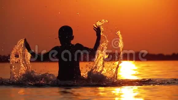 日落时分男孩的剪影举起双手创造出飞溅的水慢动作视频的预览图