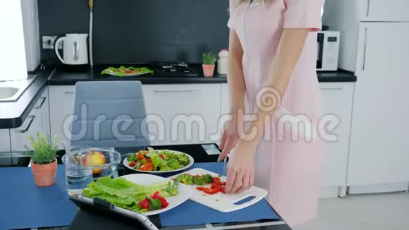 年轻女性准备蔬菜沙拉在厨房品尝甜椒并用耳机听音乐视频的预览图