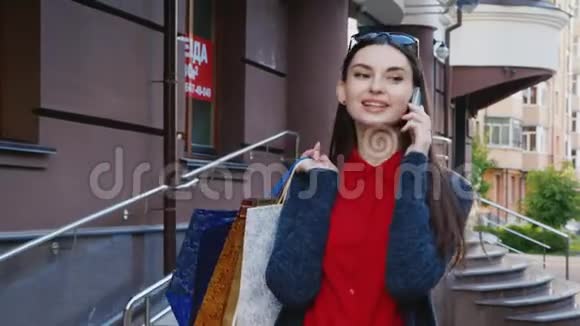 时髦的黑发女人带着购物袋在城市里走来走去在电话里交谈视频的预览图