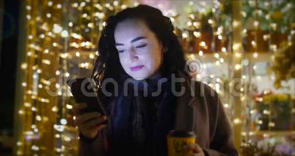 在节日的背景下迷人的女人一边用手机喝咖啡一边走在街上视频的预览图