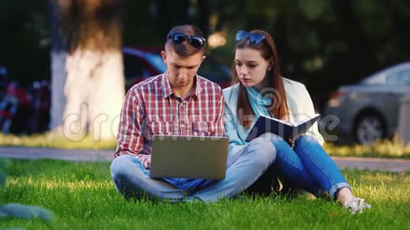 一男一女的朋友坐在公园的草地上享受着笔记本电脑在背景下人们走路视频的预览图