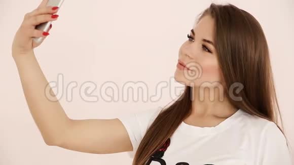现代青年漂亮的长头发长着棕色头发的欧洲女孩在白色墙壁附近的智能手机上自拍视频的预览图