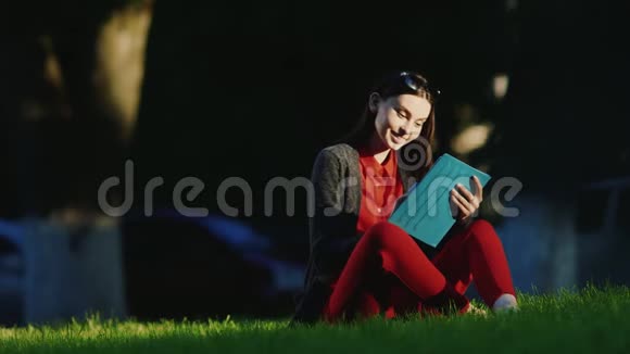 年轻的女人喜欢公园里的平板电脑坐在草地上他的脸被黄昏的太阳照亮了视频的预览图
