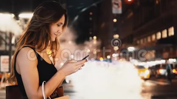 一幅年轻快乐的女人晚上站在纽约市中心的交通中使用智能手机的肖像视频的预览图