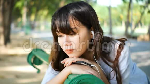 美丽的年轻时髦女郎悲伤的亚洲女人带着智能手机带着耳机听音乐坐在公园的长凳上视频的预览图