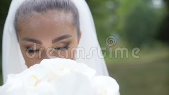 年轻迷人的黑发新娘的特写肖像闻着婚礼上的一束白色的花看着里面视频的预览图