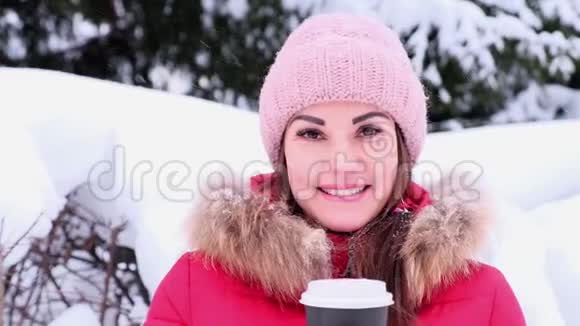 快乐的年轻女人在冬天的公园里喝着热饮料穿红色夹克衫的女性在冬天阳光明媚的日子里喝外卖咖啡视频的预览图