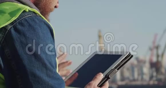 一个在黄色头盔中有胡子的肖像画工作者在海岸的背景下使用平板电脑视频的预览图