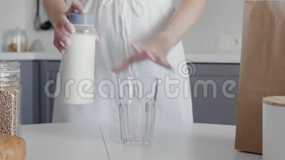 一位身穿白色连衣裙面目全非的女士将牛奶从老式瓶子倒入桌子上的玻璃杯中作为早餐视频的预览图