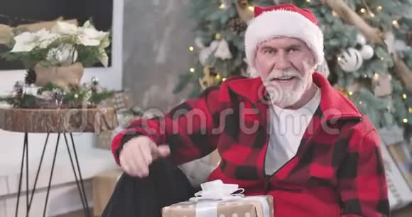 英俊的高白种人坐在新年树的背景下挥手交谈的肖像快乐圣诞老人视频的预览图