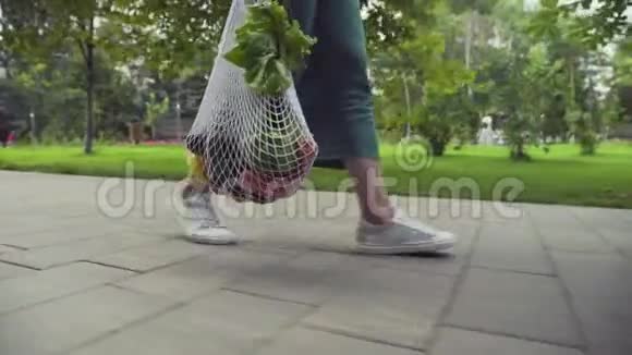 年轻女子穿着绿色裙子和白色运动鞋手里拿着带蔬菜的棉网购物袋走进来视频的预览图