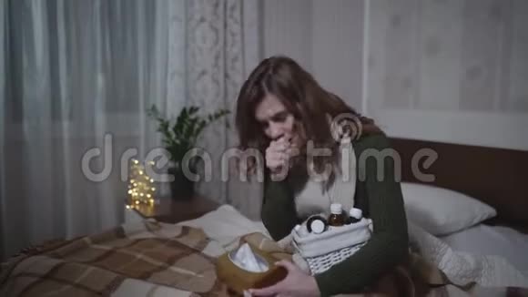 生病的女孩穿着暖和的衣服鼻塞打喷嚏和咳嗽坐在床上拿着餐巾纸和药品生病了视频的预览图