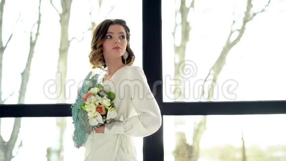 美丽的黑发新娘拿着一束花束靠近窗户头发中的精美头饰专业化妆视频的预览图