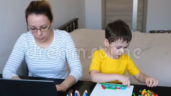 年轻的黑发母亲在她的笔记本电脑上工作她的儿子坐在桌子上在他的平板电脑上玩视频的预览图