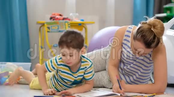 年轻快乐的母亲和小儿子躺在孩子们的房间里一起做工艺品一起画画视频的预览图