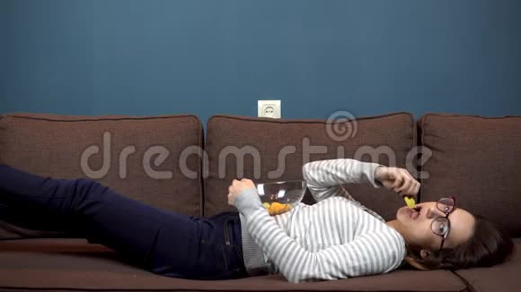 一个戴眼镜的年轻女人吃薯片看电视躺在沙发上的女孩视频的预览图