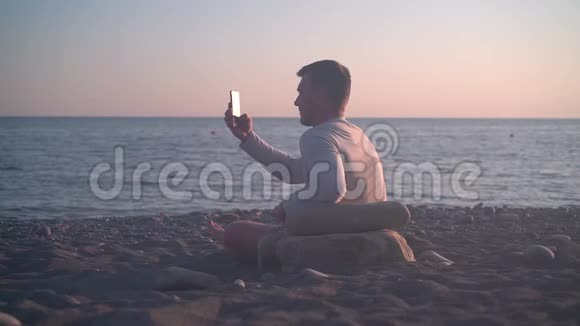 一个年轻人坐在岸边映衬着海上日落人在日落背景下在海滩上拍照视频的预览图