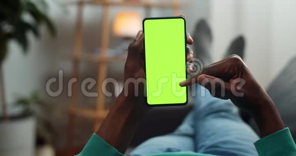 非洲男人滚动智能手机模拟显示时躺在沙发上年轻人一边上网一边看绿色视频的预览图