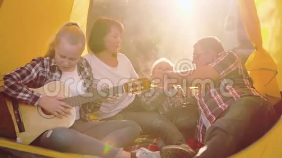 旅游家庭在阳光明媚的森林里露营帐篷里休息和演奏吉他音乐笑着一家人唱着歌视频的预览图