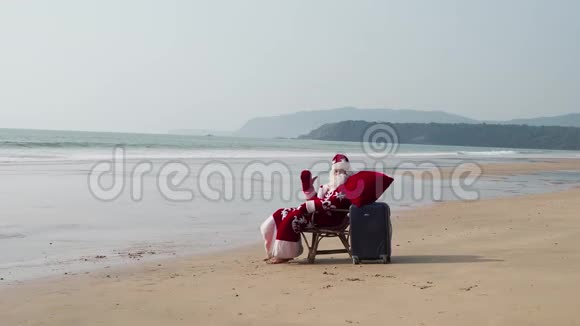 圣诞老人带着一个手提箱和一个红色的袋子坐在海边的椅子上挥动他的手视频的预览图
