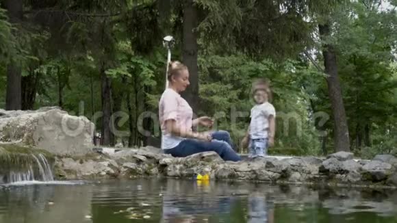 一个悲伤的小男孩坐在母亲腿上看着一个夏天在城市喷泉的湿纸船视频的预览图