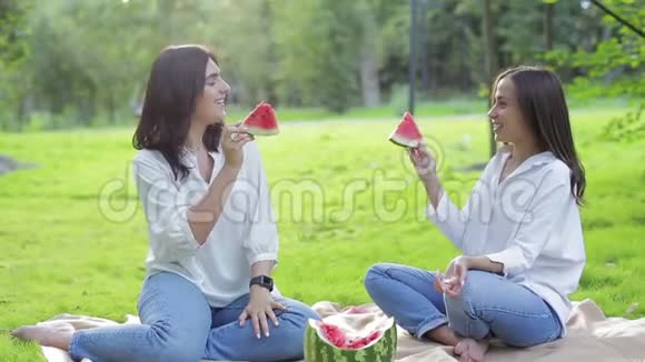 两个女孩吃着一个红色健康多汁的西瓜在公园的草地上傻笑欢快的水果野餐视频的预览图