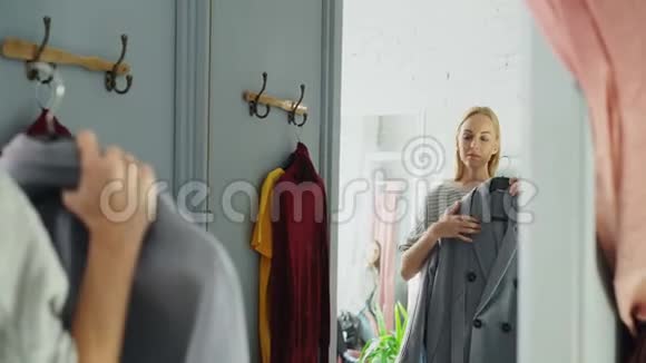 漂亮的年轻女子正在尝试优雅的外套而站在试衣间是服装精品店她在看视频的预览图