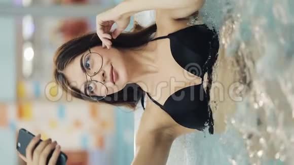 垂直视频漂亮的黑衣年轻女子在智能手机上用全新的按摩浴缸或游泳池自拍视频的预览图