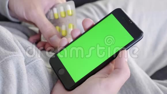 男人在家坐在沙发上拿着绿色的模拟屏幕智能手机吃药视频的预览图