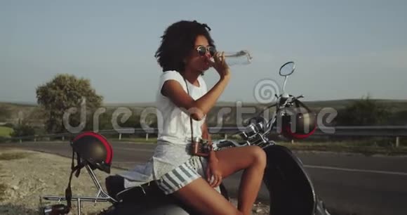 一个口渴的女孩坐在路边的摩托车上喝水4k视频的预览图