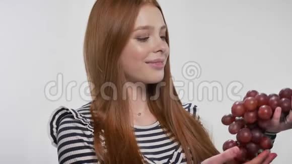 年轻的甜姜姑娘拿着葡萄吃了一个看着镜头白色背景视频的预览图