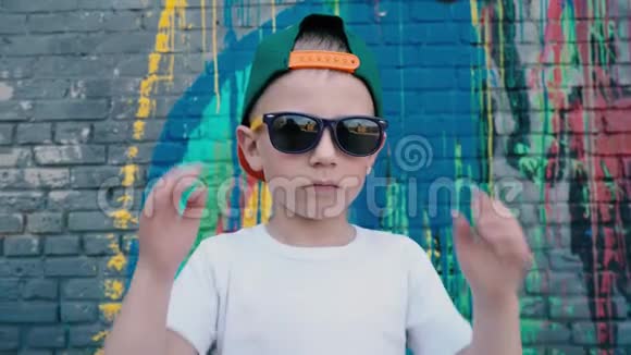 一个5岁的男孩戴着帽子戴着太阳镜在墙边的相机上涂鸦一个戴着太阳镜的孩子视频的预览图